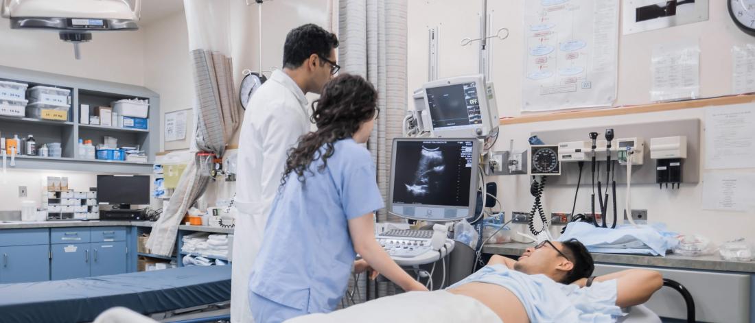 Two medical professionals look at a monitor while standing beside a patient in a bed.