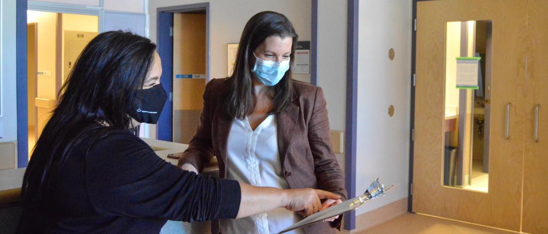 Two women stand in a hospital hallway looking at a clipboard together. 