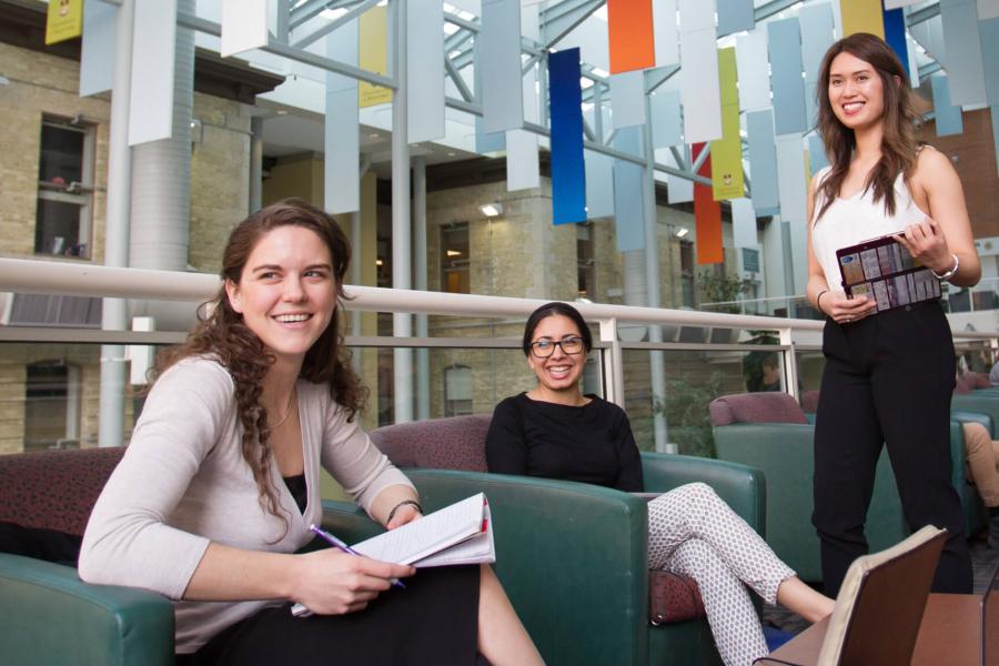 Three students smile while relaxing inside the Brodie atrium at the University of Manitoba.