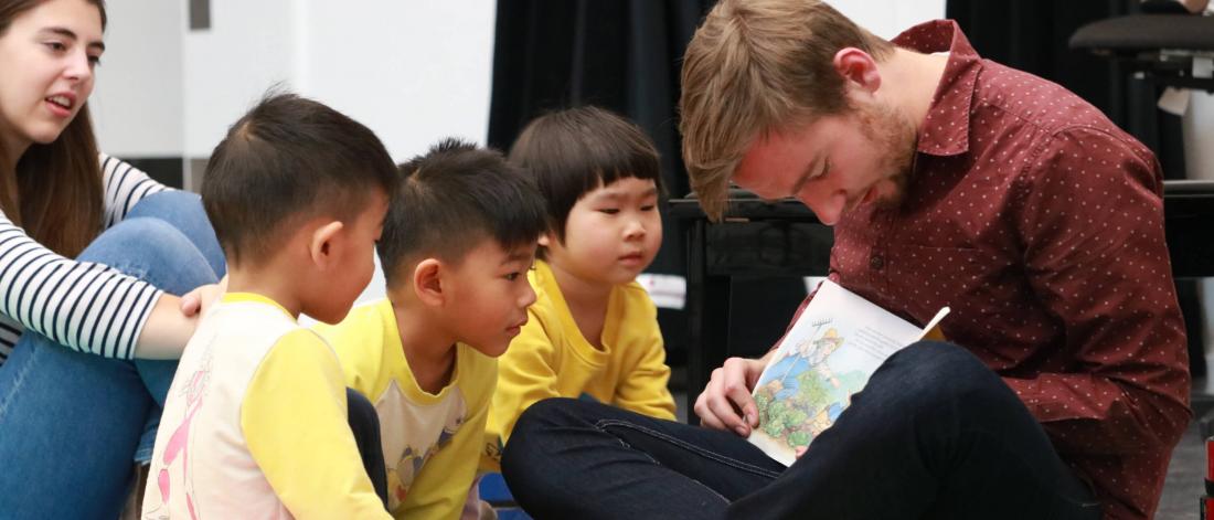 A group music lessons instructor sits on the floor and reads a book to the young participants.