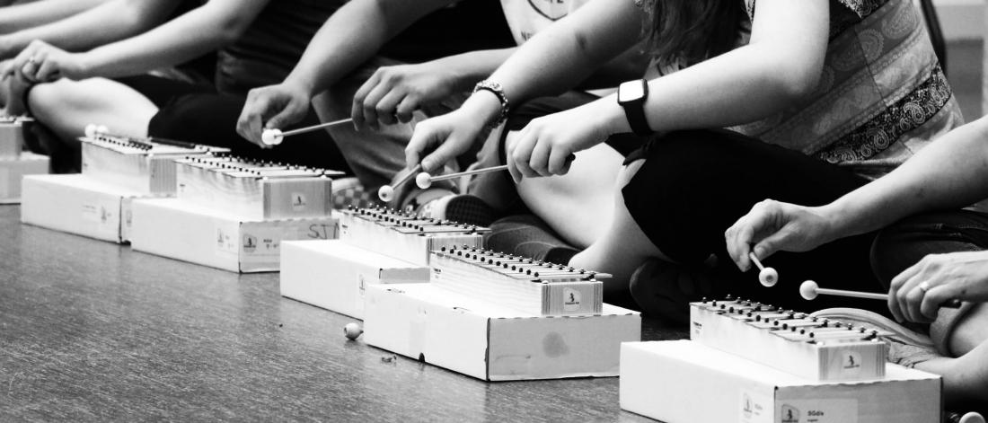 a group of people lined up sitting cross legged on the floor playing xylophones. 