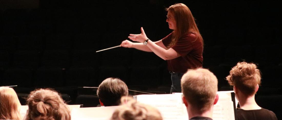 A conductor raises her baton and stretches out her arms as a wind band looks on.