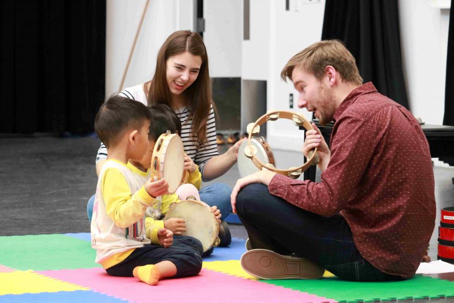 Two early childhood educators shake tambourines with toddlers.