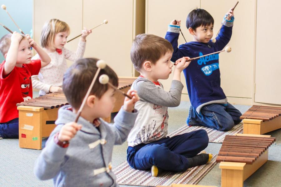 A small group of young children sit on the floor playing with xylophones. 