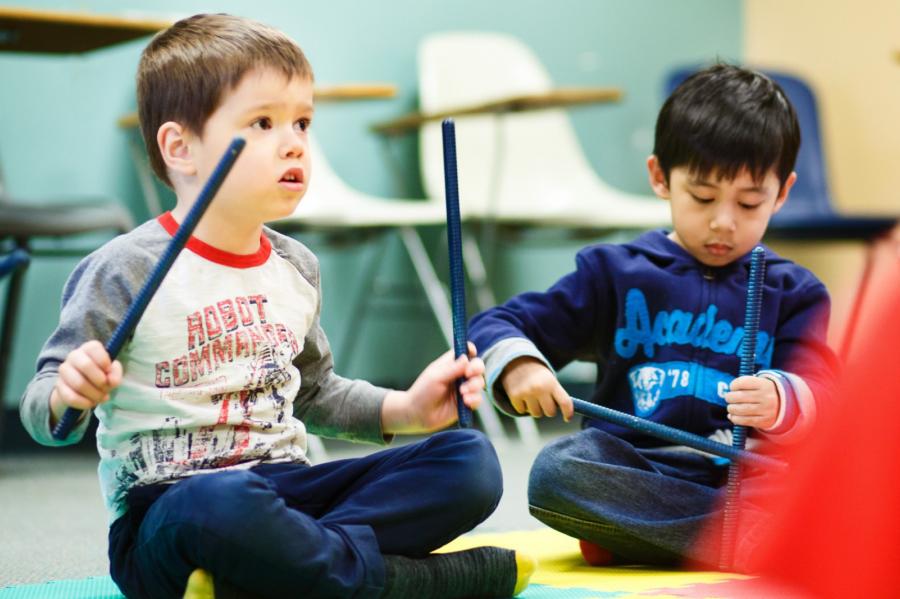 Two toddlers sit on the floor playing with drum sticks.