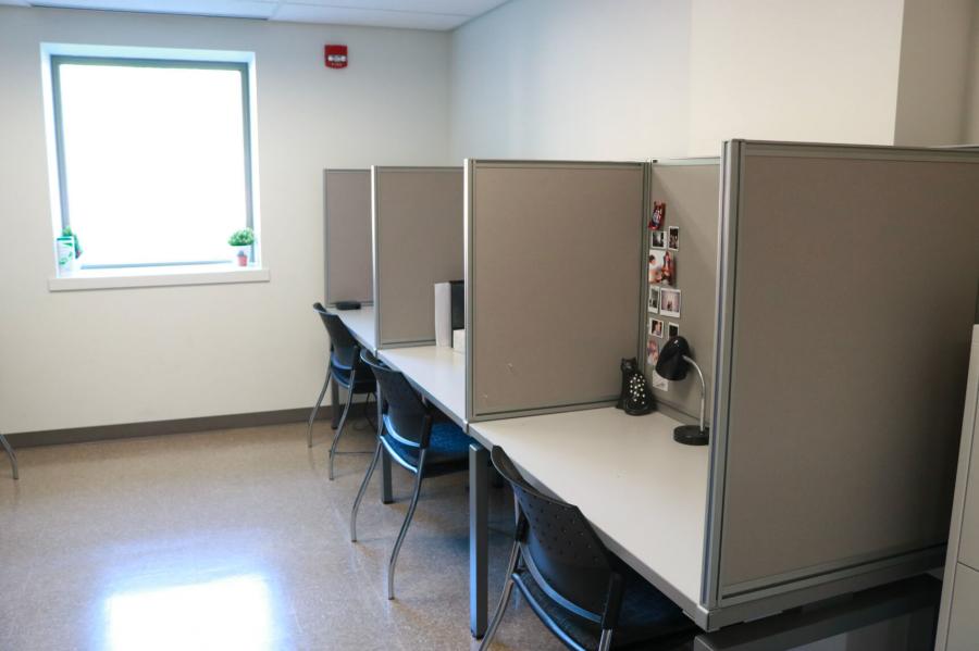 A brightly lit room with a window, and a long table along a wall partitioned off to created individual student office spaces decorated with photos and lamps. 