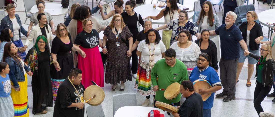 A large group of people standing in a circle, holding hands with a drum group drums in the centre of the circle.