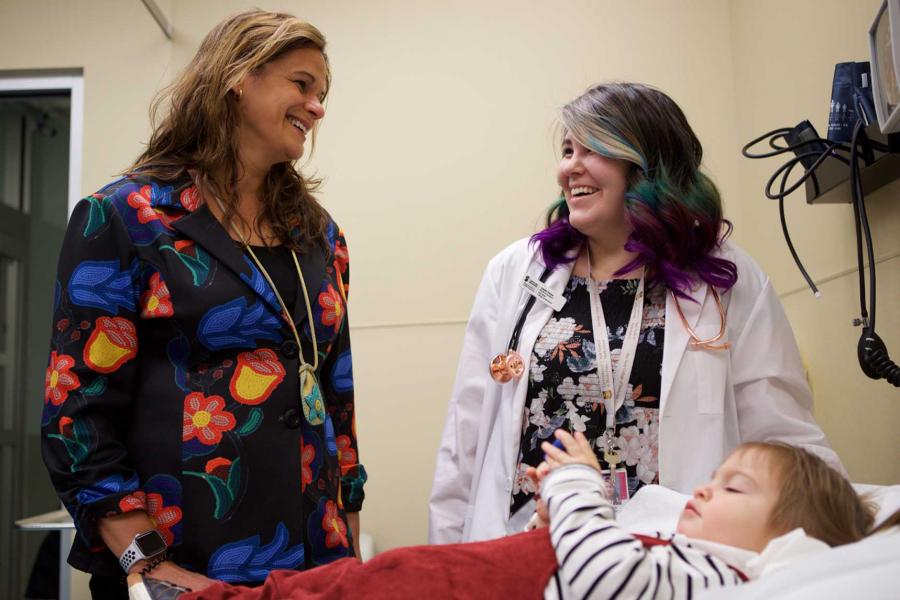 A student and a doctor share a smile while a toddler lays on an examination table.