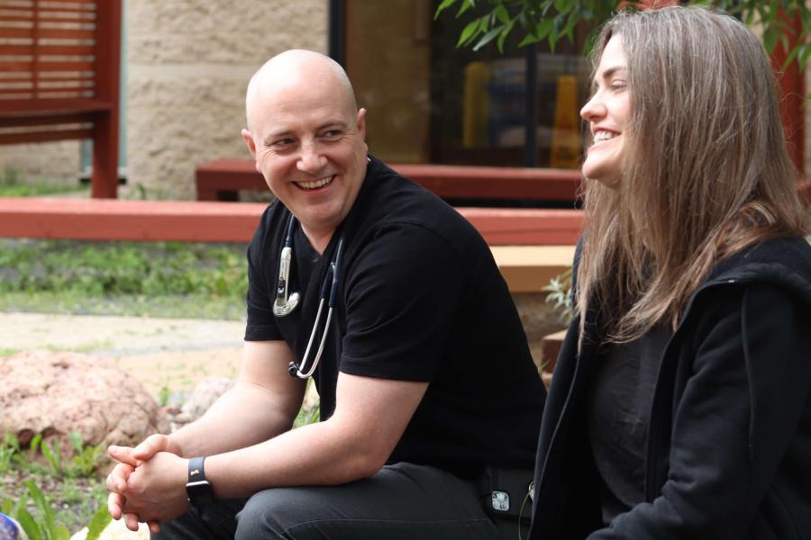 A man and woman sit on a bench in the medicine garden. Both are smiling.