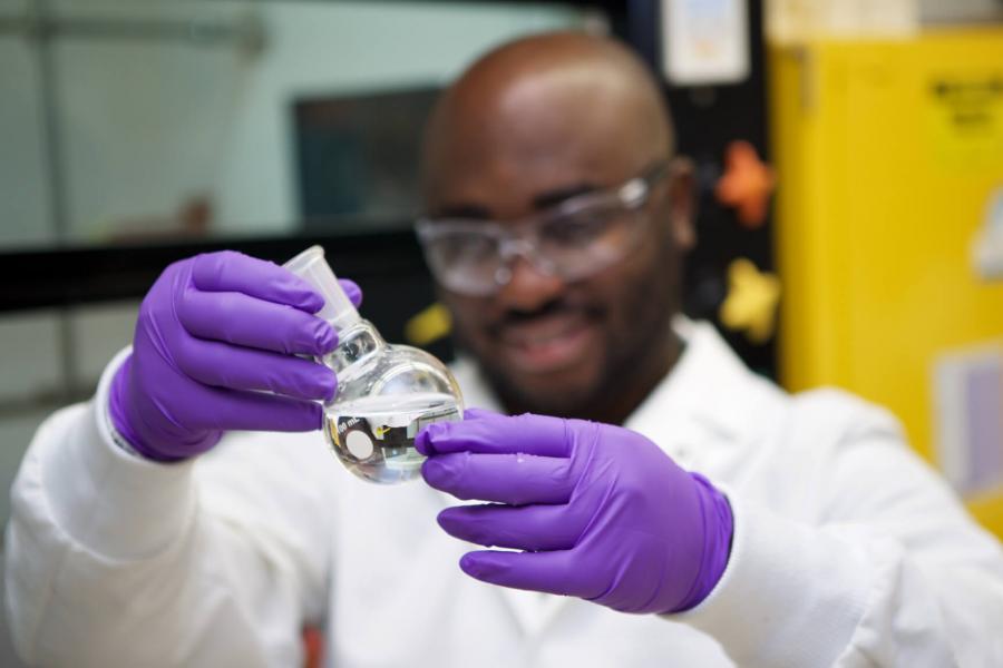 A pharmacist holds up a beaker filled with liquid to inspect it while wearing protective gloves, eyewear and a lab coat.