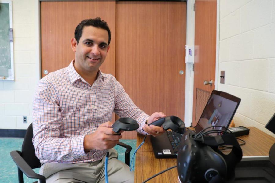 A smiling Dr. Amine Choukou sits at a table in front of a laptop holding some equipment.