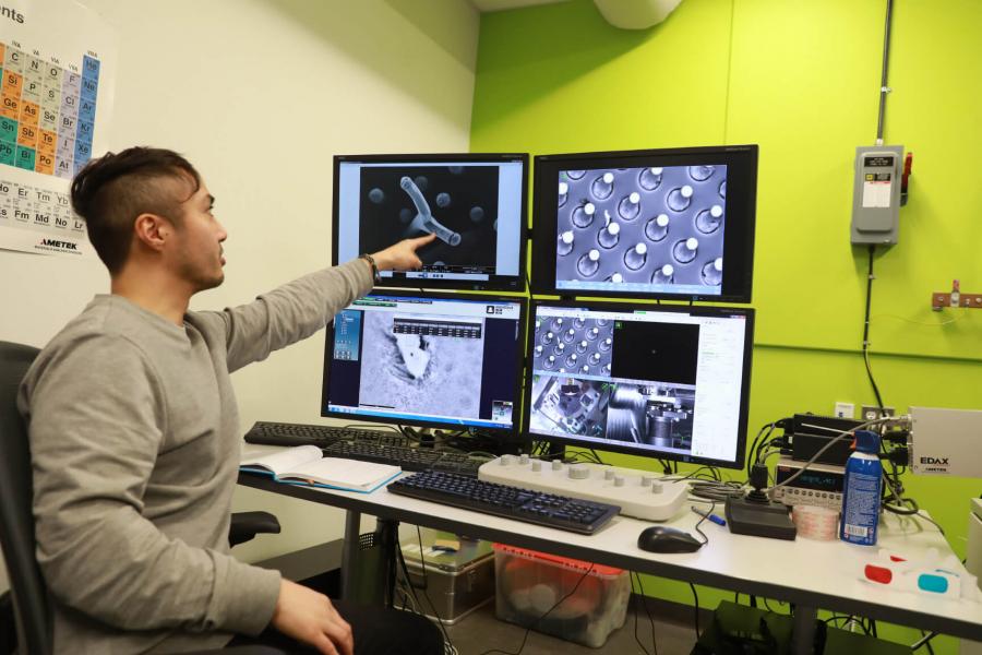 A member of the Manitoba Institute for Materials sits at a desk and points to one of four computer monitors during a demonstration.