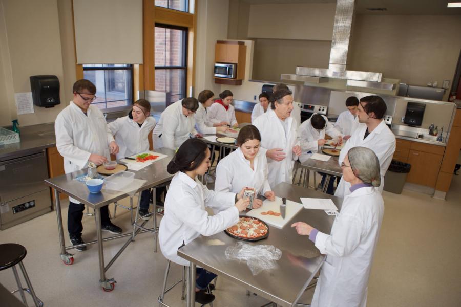 A large group of participants in a food lab wearing white coats and sitting at tables with various food items.