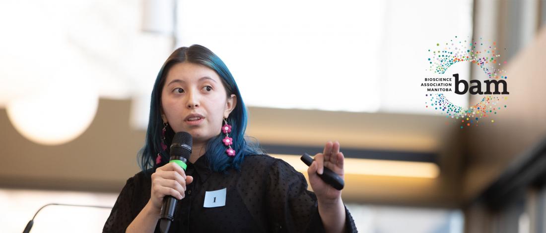 A young female student with blue hair and pink earrings in black attire holding a microphone and pointing to the right side of the photo and looking that way while presenting on stage with the Bioscience Association Manitoba (BAM) logo attached on the top right corner of the image as well.