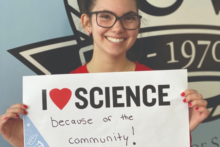 Student smiling and holding a card with the words "I love Science because of the community!".