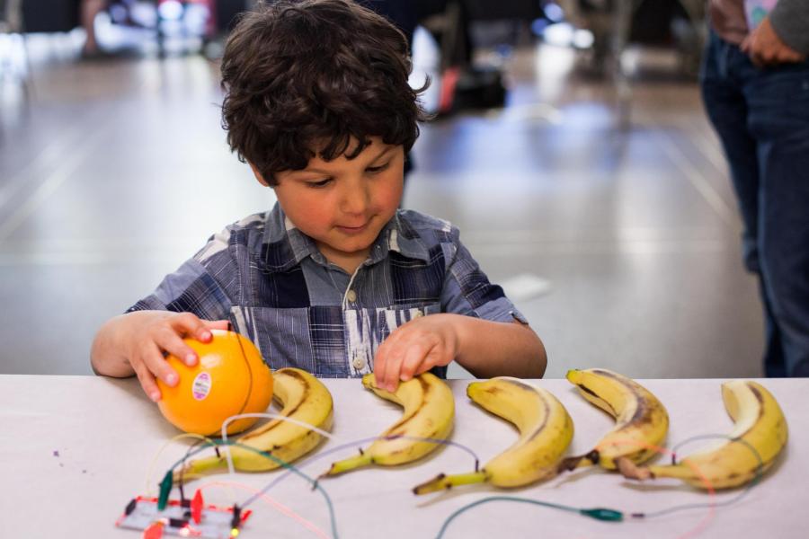 A kid touching bananas and an orange that are connected to wires.