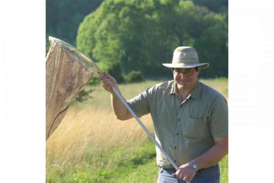 Jeffrey Marcus holding a butterfly net in a green field wearing a hat and smiling at the camera.