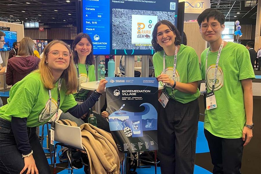 UM iGEM team of students standing and crouching in front of a TV with their logo displayed on it while holding their presentation poster all smiling and wearing green shirts with iGEM logo on them.