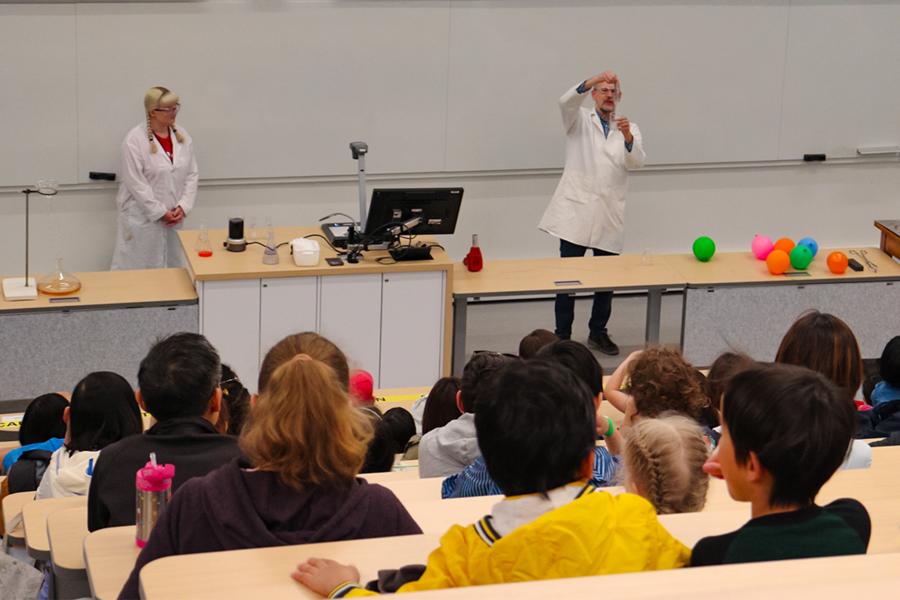 Two scientists with lab coats showing chemistry experiments to a classroom full of kids.