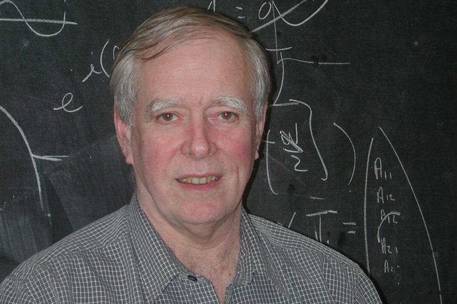 Byron Southern wearing a checkered button down shirt and smiling at the camera in front of a blackboard with equations.
