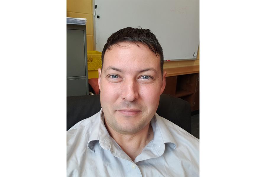 Kyle Shiells wearing a light gray button down shirt and smiling at the camera in front of a white board.