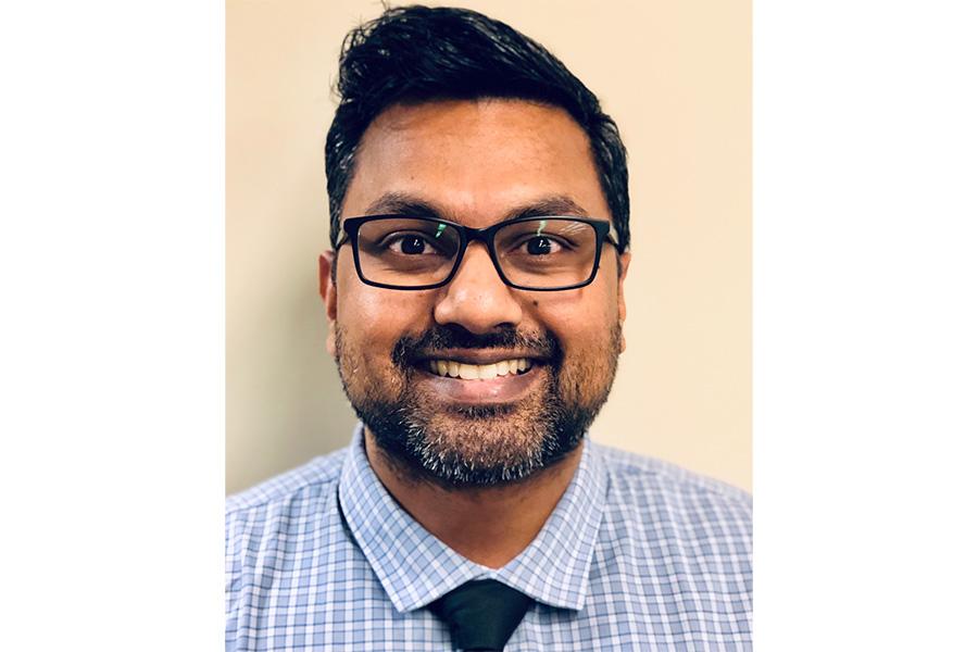 Nranjan Venugopal wearing a blue patterned button down shirt with a black tie and wearing an eyeglass smiling at the camera.
