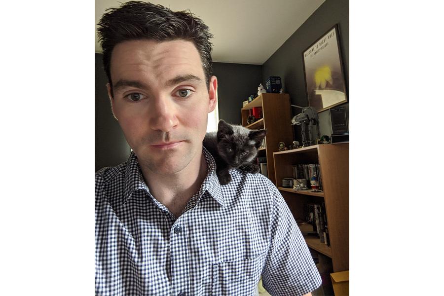 Bryan McIntosh wearing a plaid button down shirt and has a cat on the shoulder smiling at the camera.