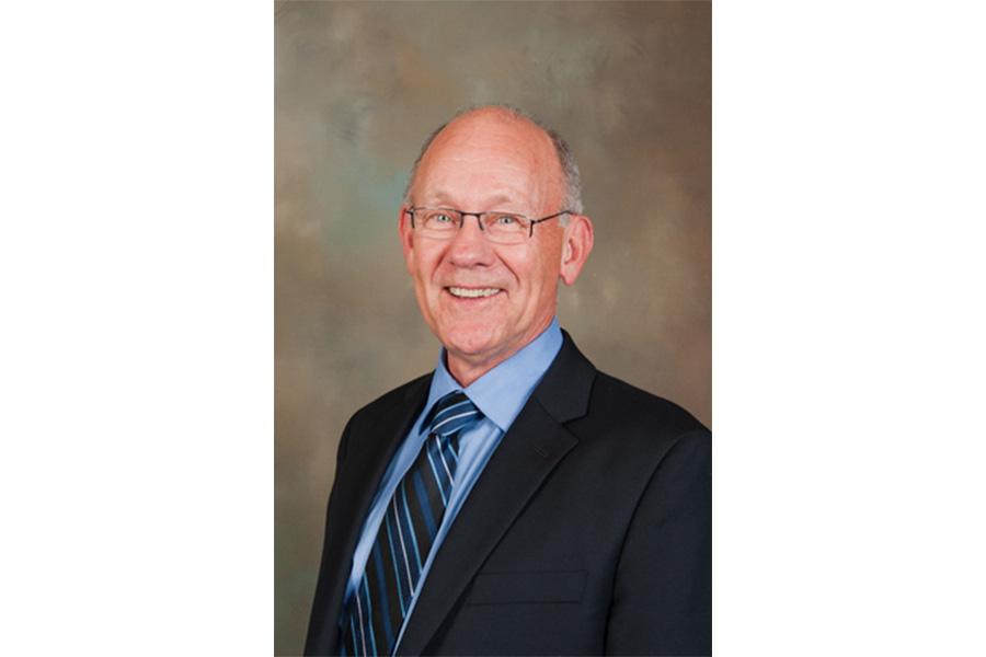 John F Brewster wearing a blue collared shirt with a coat and tie smiling at the camera.