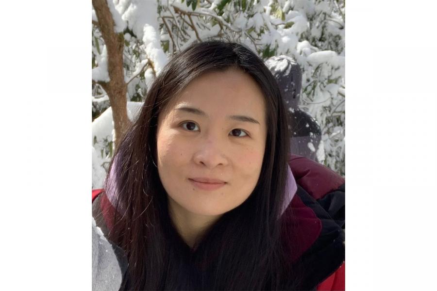 Qing Zhang wearing a purple jacket smiling at the camera with snowy trees in the background.