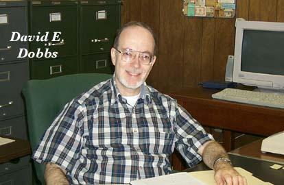 David Dobbs sitting at a desk with a plaid shirt on wearing glasses and smiling at the camera.