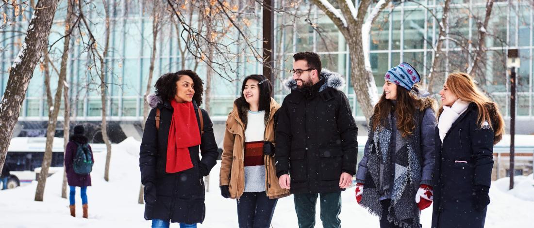 Five international students dressed in winter gear chat outside on campus, with snowbanks in the background.