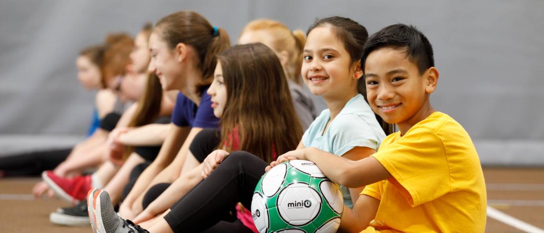 A group of children sit on the floor in a row together in a gymnasium, a young boy at the front holds a soccer ball.