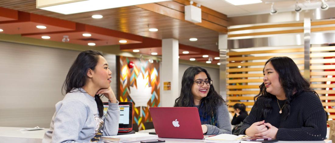 Three students sit at a table together with their laptops smiling and talking in the University Centre building.