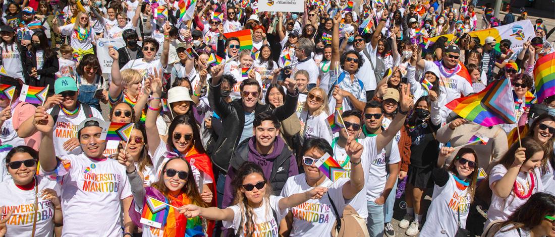 A large group of people smiling people wearing university of manitoba rainbow shirts and waving pride flags. 