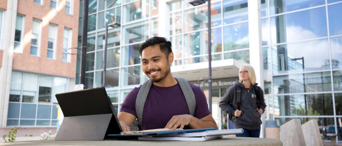 A student student sitting at a concrete picnic table sitting at a laptop in front of a building with a sign reading "Brodie Centre".