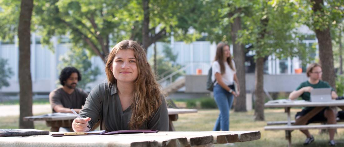 A student smiles at a picnic table on the University of Manitoba campus.