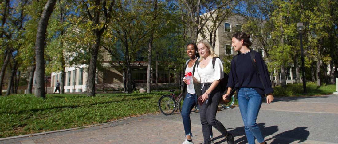 Three smiling students walking down a path on campus in front of a bike rack, building, and trees.