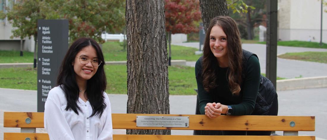 One young woman sits on an outdoor bench on Fort Garry campus; another leans up against the back of it; both are smiling.