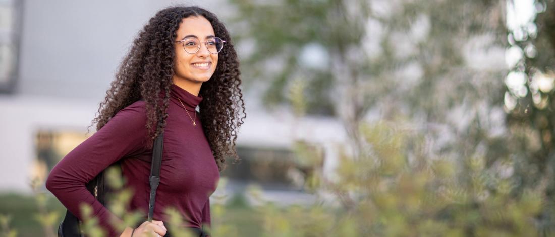 A smiling student with curly brown hair and glasses, wearing a backpack, with foliage in the background.