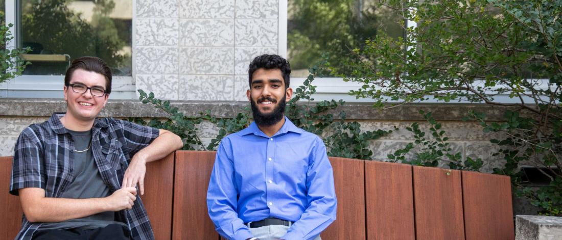 University of Manitoba students Cody McDonald and Simar Ubhi sitting on a bench on campus, smiling.