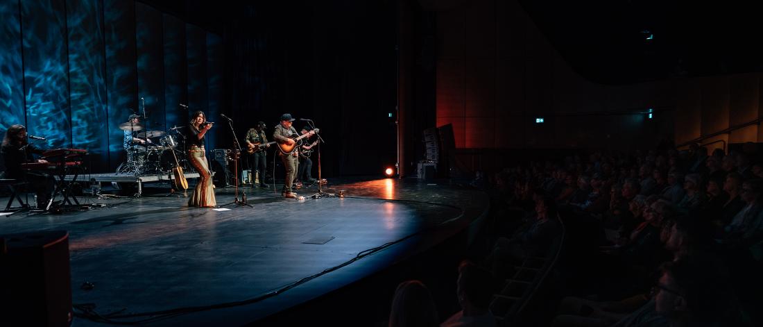 three musicians standing on stage to the left of the image, playing guitar to an audience situated on the right