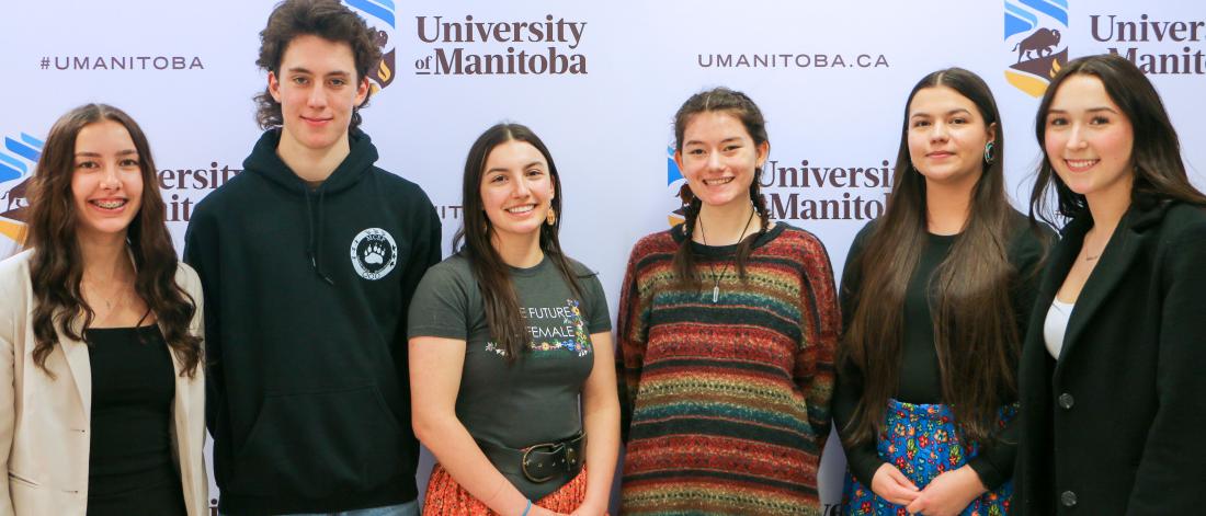 Six students in a row in front of a UM logo backdrop.