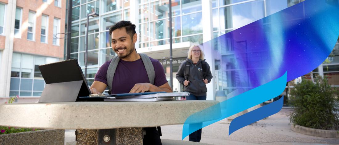 A student sits and smiles at a table as another student walks past near the Brodie building on the Bannatyne campus.