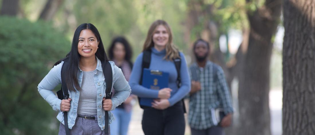 A student smiles and walks along a treed sidewalk.