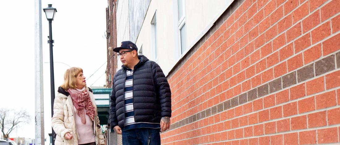 Associate professor Marleny Bonnycastle in a white coat and Henry McKay who is a past undergraduate research award recipient, wearing a black coat, walk and talk on a sidewalk beside a brick building in downtown Winnipeg.