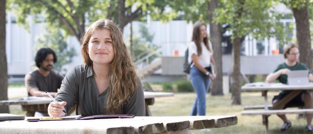 A student sits at a picnic table and smiles with her book out.