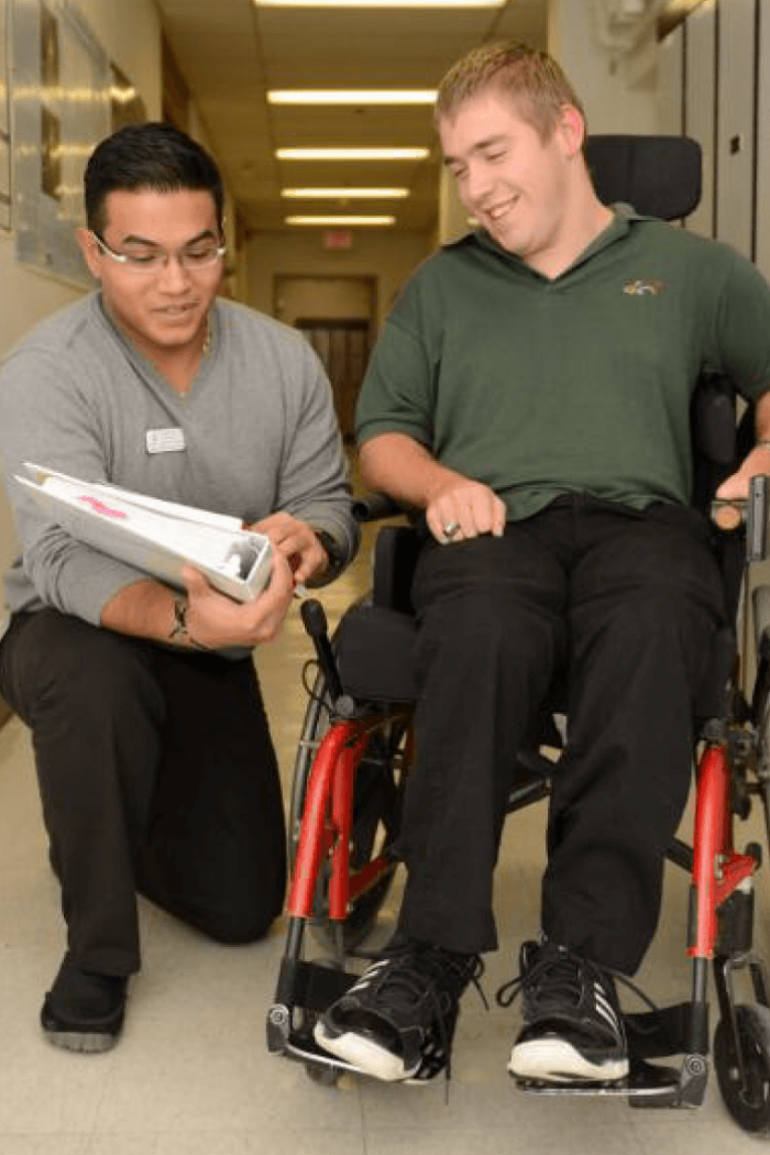 A student kneels down on one knee as he speaks to a person who is using a wheelchair and points to papers he is holding. 