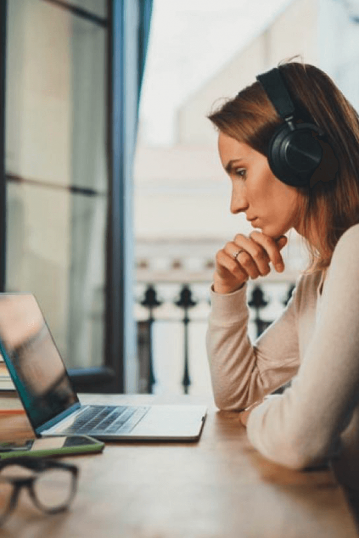 A woman wearing headphones seated at a table, leans on her elbows as she watches her laptop monitor.