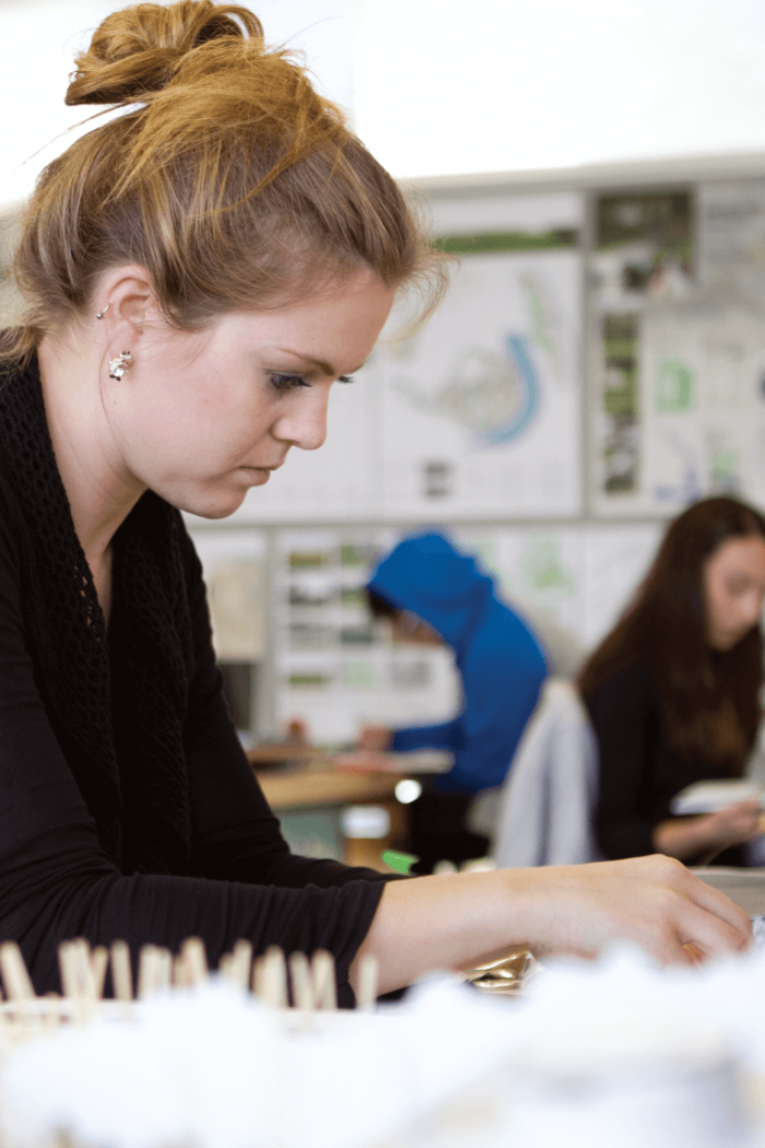 A students works on a project at a desk. Several other students can be seen working in the background.