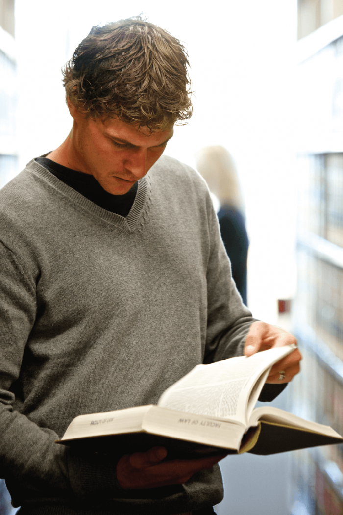 A student reads a book while standing at a bookshelf in a library.